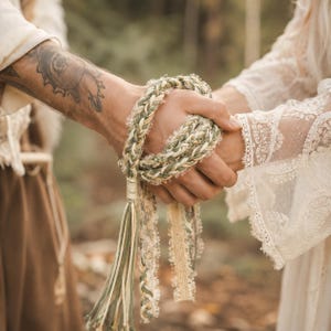 Puede incluir: Un cordón de ceremonia de mano con hebras trenzadas en verde salvia, crema y blanco, entrelazadas con cinta de encaje delicada. El cordón termina en una borla decorativa, mostrando una mezcla de texturas y tonos neutros suaves.
