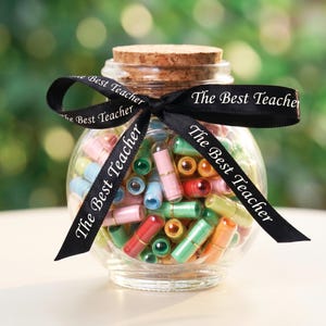 Può includere: Un barattolo di vetro trasparente pieno di messaggi di carta arrotolata colorata, sormontato da un tappo di sughero e legato con un nastro nero. Il nastro riporta la scritta "The Best Teacher" in bianco. Il barattolo è posto su uno sfondo verde sfocato.
