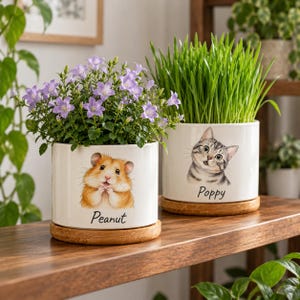Könnte beinhalten: Zwei weiße zylindrische Keramik-Pflanzgefäße mit Holzuntersetzern. Eines zeigt einen Hamster mit dem Schriftzug Peanut und violette Glockenblumen. Das andere zeigt eine Katze mit dem Schriftzug Poppy und grünes Gras.