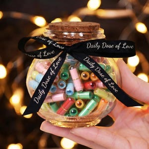 Könnte beinhalten: Ein klares Glasgefäß gefüllt mit bunten Kapseln, gebunden mit einem schwarzen Band mit der Aufschrift "Daily Dose of Love". Das Glas hat einen Korkdeckel und ist mit aufgerollten Papiernachrichten in verschiedenen Farben gefüllt.