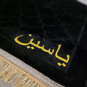 May include: Black prayer mat with gold Arabic script. The mat has a textured pattern and is trimmed with a gold fringe and a silver border. The text on the mat reads "Yaseen".