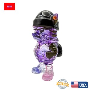 May include: A translucent purple figurine of a cartoon character wearing a black hat and jacket. The figurine has a unique design with visible internal structures. The base of the figurine is a mix of blue and pink. The word "HBOH" is in a red box.