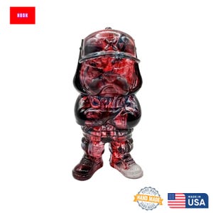 May include: A red and black bulldog figurine wearing a hat. The figurine has a stern expression and crossed arms. The figurine is hand made in the USA.