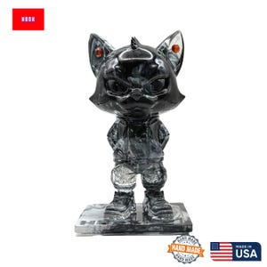 May include: A black and grey cat figurine with a stern expression, wearing a jacket and trousers. The figurine has red accents on its ears and is standing on a rectangular base. The text "HBOH" is in a red box in the upper left corner.