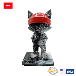 May include: A black and grey cat figurine wearing a red beanie and jacket. The figurine stands on a black base with "HBOK" text. The figurine is hand made in the USA.