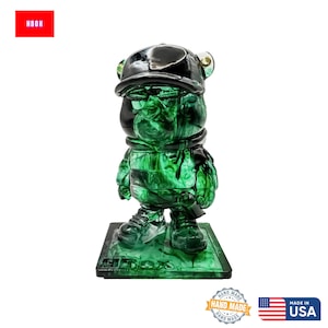 May include: A green resin bear figurine wearing a black hat and scarf. The figurine stands on a green base with the text "HBOX". The figurine has a glossy finish and is made in the USA.