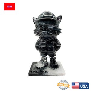 May include: A black cat figurine wearing a hat and jacket, standing on a rectangular base. The figurine has a glossy finish and is detailed with visible ears and boots. The base has the text "HBOK" and "HAND MADE IN USA" is printed on the bottom.