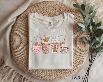 Op de afbeelding: Ivoor kleurig t-shirt met een feestelijk ontwerp van vier mokken warme chocolademelk, elk versierd met peperkoekmannetjes en slagroom. Het ontwerp bevat sneeuwvlokken en sterren, perfect voor de feestdagen.