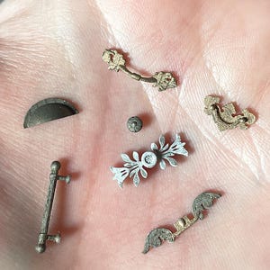 Puede incluir: Una comparación lado a lado de herrajes en miniatura sin pintar y pintados. Los herrajes sin pintar son de color gris claro, mientras que los herrajes pintados son de una variedad de colores metálicos, incluyendo oro, plata y bronce. Los herrajes incluyen tiradores de cajones, pomos de armario y manijas de puertas. La imagen incluye el texto "archivo digital", "antes de pintar" y "después de pintar".