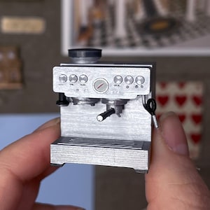 May include: A miniature silver espresso machine with a black handle and dials. The tiny appliance is held in a hand, with various miniature artworks and a "digital file" banner in the background. The espresso machine is a detailed replica.