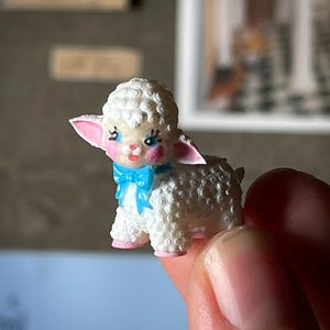 May include: A miniature white lamb figurine with a curly wool texture, pink ears and feet, and a blue bow. The lamb has a sweet expression with rosy cheeks and is held in a person's hand.