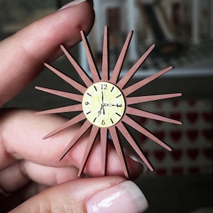 May include: A miniature, starburst-shaped clock with a light brown frame and a cream-colored clock face. The clock hands point to approximately 12:05. The words "digital file" are in a banner in the upper right corner. The clock is held in a person's hand.