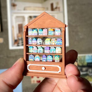 Może przedstawiać: Miniaturowa brązowa drewniana szafka z dwuspadowym dachem, wypełniona kolorowymi małymi domkami. Szafka ma szufladę i jest trzymana w dłoni osoby. Tło zawiera dzieła sztuki i słowa "digital file".