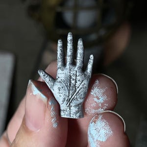May include: A miniature, white and gray hand sculpture with intricate line details, held between fingers. The hand is decorated with symbols and lines, resembling a palmistry chart. The image includes a banner that reads "digital file".