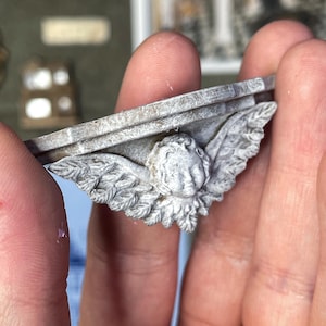 Può includere: Una piccola mensola decorativa grigia con un volto d'angelo e ali scolpite sul davanti. La mensola è tenuta in una mano. Le parole "digital file" sono in uno striscione in alto a sinistra. Altre opere d'arte in miniatura sono sullo sfondo.