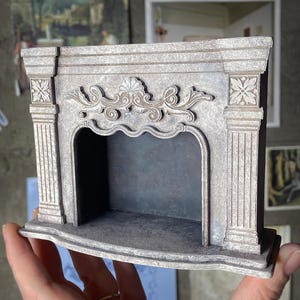 Puede incluir: Una chimenea gris en miniatura con detalles ornamentados, que incluyen columnas y tallas florales. Las palabras "digital file" están en una pancarta en la esquina superior izquierda. La chimenea se sostiene en una mano.