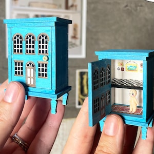 May include: Two miniature teal dollhouse cabinets, one open and one closed. The closed cabinet has arched windows and a small door. The open cabinet reveals a small interior with a rug and a figure. The cabinets are labeled "digital file".