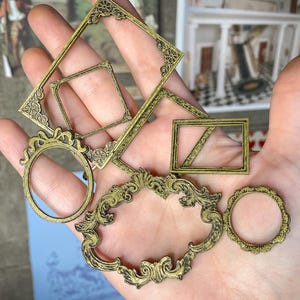 May include: A hand holds several antique brass-colored decorative frames in various shapes, including rectangular, square, and oval. The frames have ornate, detailed designs. The image includes the text "digital file" in a banner.