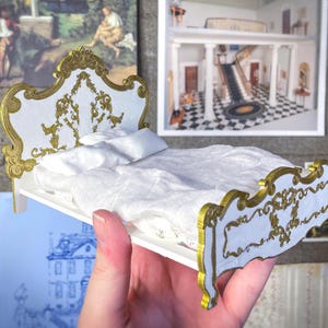 Puede incluir: Cama blanca en miniatura con adornos dorados y detalles ornamentados. El cabecero y el piecero presentan un diseño de volutas. La cama está hecha con ropa de cama blanca y una pequeña almohada. La imagen incluye el texto "digital file".