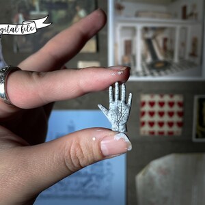 May include: A miniature, weathered-looking hand sculpture, held between fingers. The tiny hand is gray with etched lines, suggesting a palmistry design. The image includes the text "digital file" on a banner.