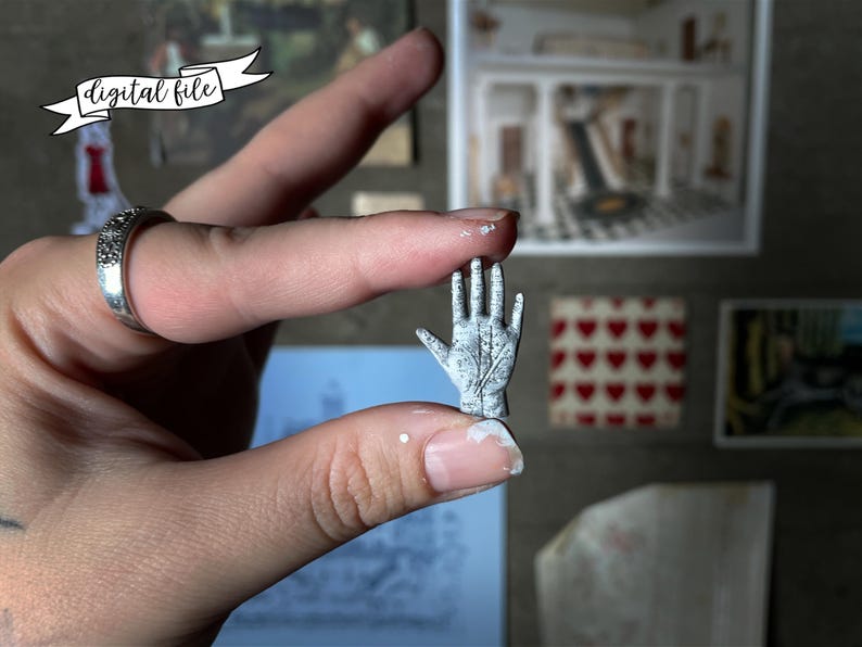 May include: A miniature, weathered-looking hand sculpture, held between fingers. The tiny hand is gray with etched lines, suggesting a palmistry design. The image includes the text "digital file" on a banner.