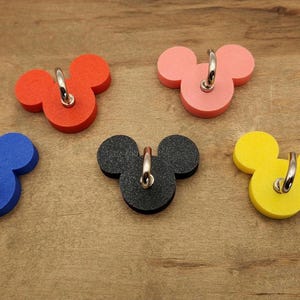 Könnte beinhalten: Fünf farbige Mickey Mouse-förmige Wandhaken in Rot, Rosa, Blau, Schwarz und Gelb. Jeder Haken hat einen silberfarbenen Metallring zum Aufhängen. Die Haken sind auf einer Holzoberfläche angeordnet.
