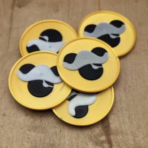 Op de afbeelding: Een verzameling gele, ronde onderzetters met een zwart en grijs ontwerp. De onderzetters hebben een verhoogde, getextureerde rand en een gestileerd ontwerp in het midden. Het ontwerp lijkt op een cartoonachtig karakter. De onderzetters zijn gestapeld op een houten oppervlak.