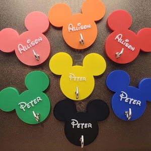 May include: A collection of colorful, Mickey Mouse-shaped wall hooks. Each hook is a different color, including pink, orange, red, yellow, green, blue, and black. Each hook has a name written on it in white script.