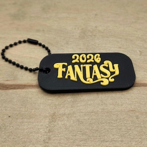 May include: A black tag with the word "FANTASY" in yellow, along with the year "2026". The tag is attached to a black ball chain. The tag is on a wooden surface.