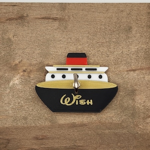 May include: A black, gold, and white ship-shaped keychain with the word "Wish" in gold script. The keychain has a red and black smokestack and a silver key ring. The background is a wooden surface.