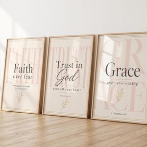 May include: Three rectangular wall art prints in light wood frames. The first reads Faith over fear, the second Trust in God with all your heart, and the third Grace over everything. Each print features soft pink text, black lettering, and a small leaf icon.