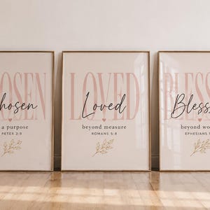 Puede incluir: Tres láminas de pared enmarcadas con fondo beige claro y texto rosa. Los carteles muestran las palabras Chosen, Loved y Blessed en fuente rosa grande, con texto en cursiva negra más pequeño e ilustraciones botánicas de líneas doradas.