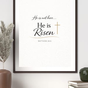 Puede incluir: Impresión enmarcada con las palabras "He is not here... He is Risen" en escritura elegante y una cruz dorada. El texto "Matthew 28:6" está debajo. La impresión está en un marco marrón oscuro, exhibida en un estante de color claro con elementos decorativos.