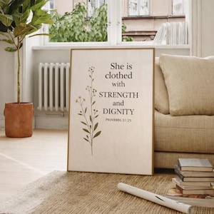 Puede incluir: Una impresión enmarcada con el texto "She is clothed with STRENGTH and DIGNITY PROVERBS 31:25" en fuente negra. La impresión tiene un fondo beige con una ilustración botánica. El marco es de color marrón claro. La impresión se muestra sobre una alfombra.