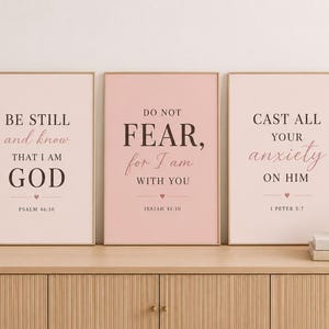 Puede incluir: Tres láminas rectangulares en marcos de color rosa claro con texto en negro y rosa. Las láminas muestran versículos religiosos: Be still and know that I am God, Do not fear for I am with you y Cast all your anxiety on him con corazones.