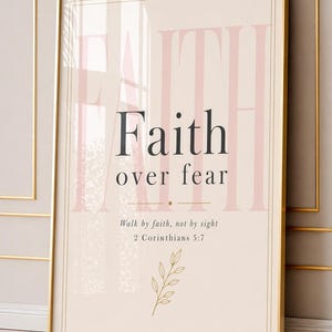 Puede incluir: Un cuadro rectangular con marco dorado fino que presenta un fondo crema con el texto grande en rosa pálido FAITH, el texto negro Faith over fear, el versículo Walk by faith, not by sight 2 Corinthians 5:7 y una hoja en dibujo lineal dorado.