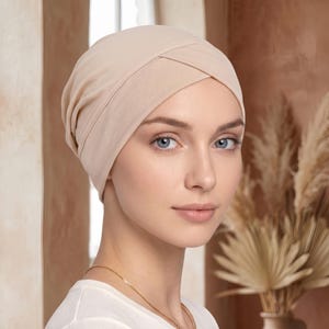 May include: A beige turban-style head covering made of soft fabric. The design features a smooth, fitted crown with a distinct crisscross detail across the front forehead area, providing a secure and comfortable fit around the head.