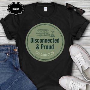 May include: Black t-shirt with a green and white graphic that reads "Disconnected & Proud Off Grid Club." The design features a camper and a campfire. The shirt is paired with blue jeans and white sneakers.