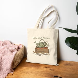 May include: Cream-colored tote bag featuring a gardening illustration with a watering can, potted plants, and gardening tools. The text "Grow What You Love" is printed above the illustration. The bag has long handles.
