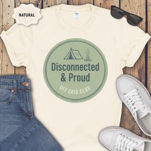 May include: Cream-colored t-shirt with a circular graphic that reads "Disconnected & Proud Off Grid Club." The graphic features a tent and campfire illustration in green. The shirt is paired with blue jeans, white sneakers, and sunglasses.