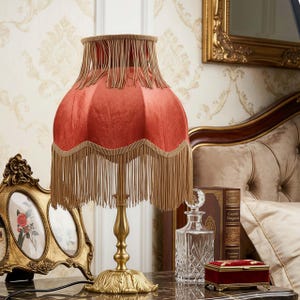May include: A vintage-style table lamp with a coral-colored shade and gold fringe. The lamp has a gold-colored base and stem. The lamp is on a table with other decorative items, including a framed picture and a crystal decanter.