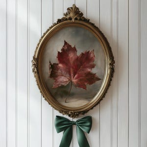 May include: An oval-shaped wall art piece featuring a painting of a single deep red maple leaf against a muted grey background. The artwork is encased in an ornate, antique gold-coloured frame with a dark green ribbon bow attached at the bottom.