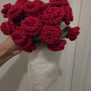 Puede incluir: Un ramo hecho a mano de rosas rojas de ganchillo. Las rosas están agrupadas, con hojas verdes visibles. El ramo se sostiene en un jarrón blanco tejido.