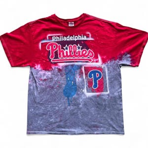 Puede incluir: Camiseta teñida con tinte rojo y gris con el logotipo de los Filis de Filadelfia. La camiseta muestra el nombre y el logotipo del equipo en blanco y rojo. También se ve una silueta azul y la letra "P".