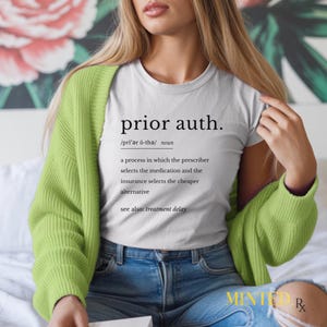 以下が含まれることがあります： 「prior auth.」の文字と定義が書かれた白いTシャツ。定義は、処方医が薬を選択し、保険会社がより安価な代替品を選択するプロセスについて説明しています。シャツは、グリーンのカーディガンとジーンズと一緒に着用されています。