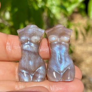 Pode incluir: Duas pequenas esculturas iridescentes da forma feminina. As esculturas s&atilde;o de cor perolada com tons azuis e roxos. As figuras s&atilde;o lisas e detalhadas, com seios definidos e uma sugest&atilde;o da parte inferior do corpo. O fundo &eacute; verde desfocado.