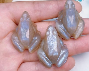 Pode incluir: Três figuras de rã esculpidas em cinza claro com tons iridescentes azuis e roxos. As rãs estão em posição sentada com detalhes. As figuras são seguradas em uma mão.