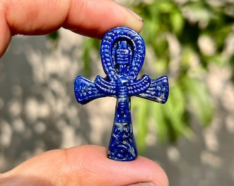 Pode incluir: Um pequeno pingente de lápis-lazúli azul profundo esculpido no formato de um símbolo egípcio ankh. A pedra apresenta manchas naturais brancas e douradas em toda a superfície, com detalhes gravados intrincados representando um pássaro no centro.