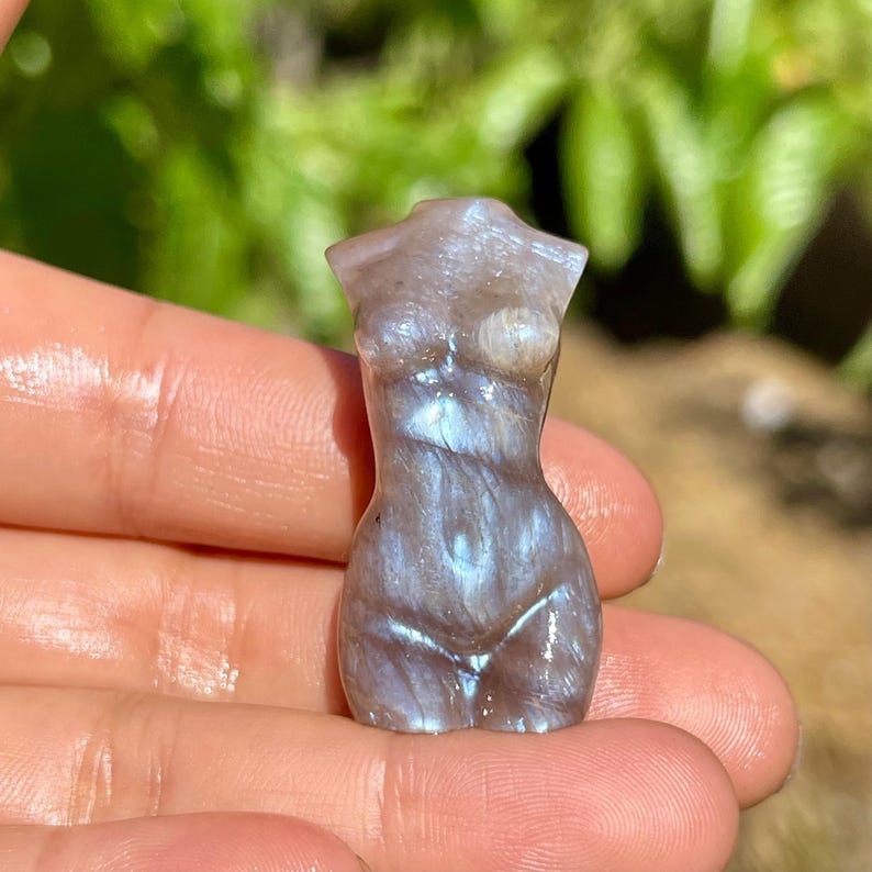 Pode incluir: Pequena estatueta esculpida em pedra preciosa representando uma forma feminina. A escultura &eacute; cinza perolado com flashes iridescentes azuis e prateados. A figura &eacute; segurada em uma m&atilde;o contra um fundo verde desfocado.