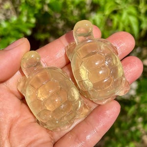 以下が含まれることがあります： 滑らかで磨かれた素材から彫られた、半透明の淡い黄色の小さなカメの置物2体。それぞれのカメは、目に見える六角形の模様がある詳細な甲羅、丸みを帯びた頭、4つのひれを備えており、透明感のあるガラスのような質感を特徴としています。